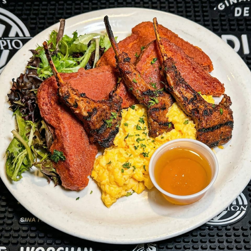 Grilled lamb chops, red velvet pancakes, scrambled eggs, and a side of syrup on a white plate.