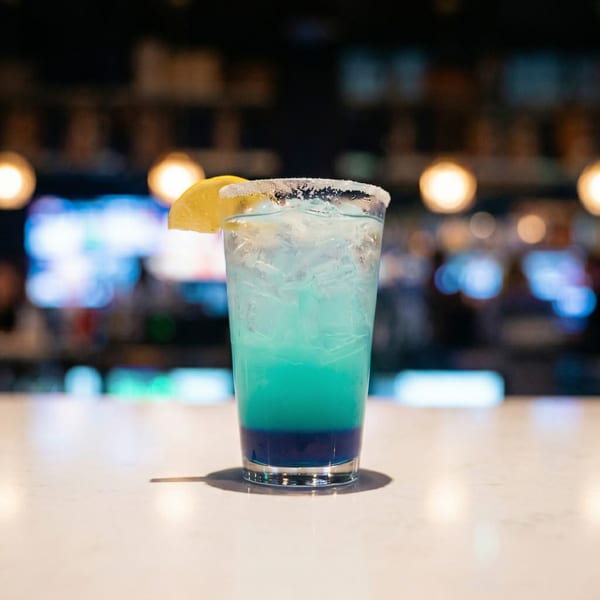 A vibrant blue and green layered cocktail with a sugared rim and lemon slice sits on a bar.